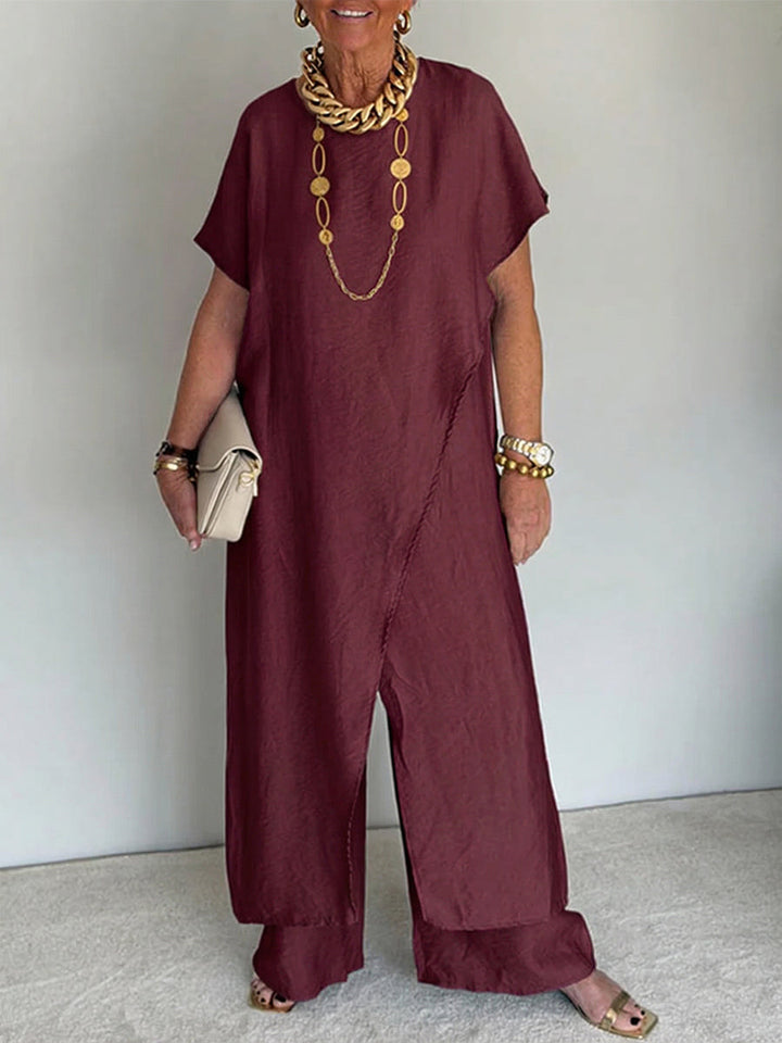 Person wearing a burgundy flowing set with short sleeves and wide-leg pants, accessorized with gold jewelry and holding a beige clutch in a studio setting.