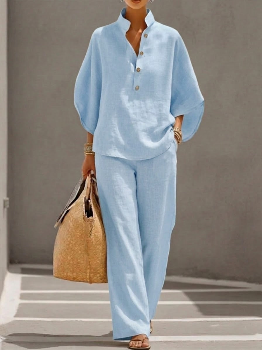Woman wearing a light blue two-piece set with a high collar and buttons, carrying a woven bag, walking in a minimalist urban setting.