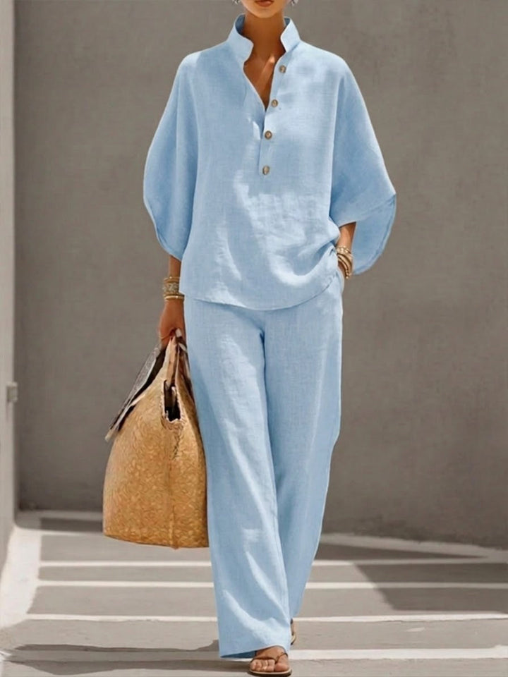 Woman wearing a light blue two-piece set with a high collar and buttons, carrying a woven bag, walking in a minimalist urban setting.