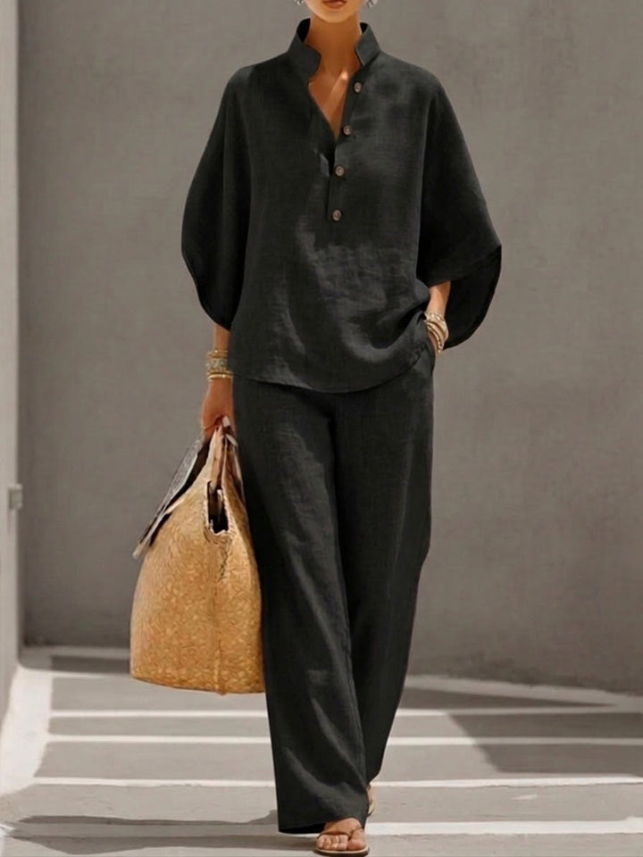 Woman wearing a black two-piece set with a high collar and buttons, carrying a woven bag, walking in a minimalist urban setting.