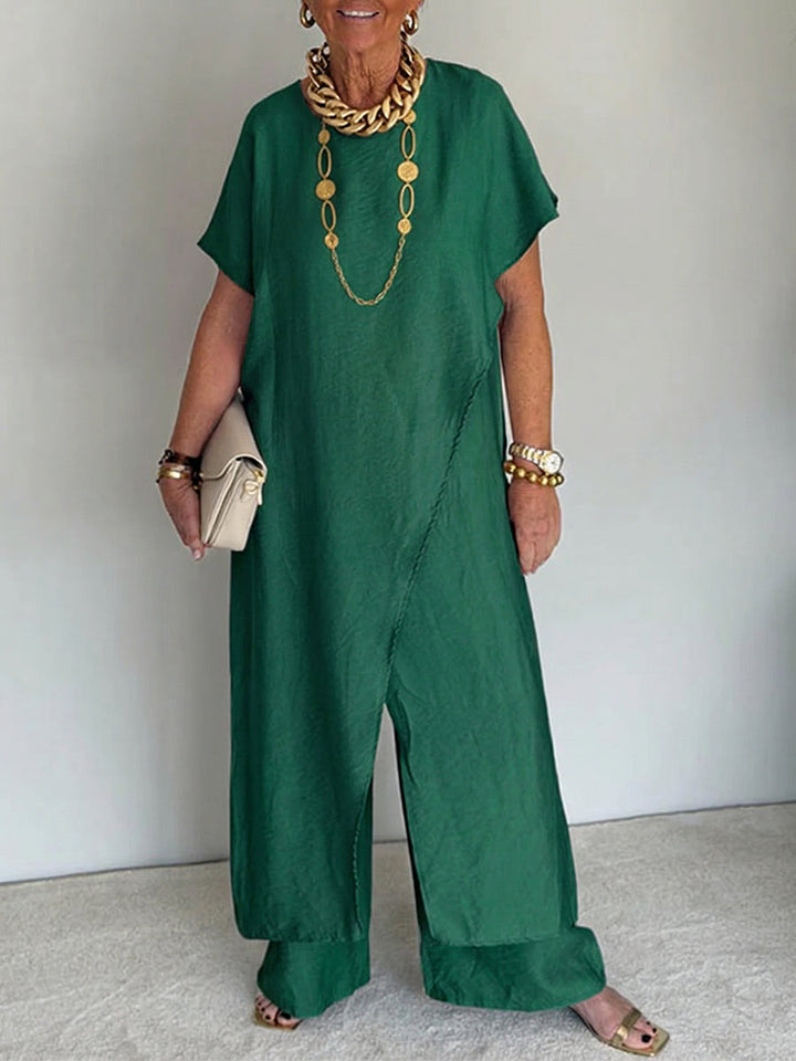 Person wearing a deep green flowing set with short sleeves and wide-leg pants, accessorized with gold jewelry and holding a beige clutch in a studio setting.