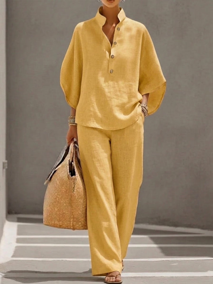 Woman wearing a yellow two-piece set with a high collar and buttons, carrying a woven bag, walking in a minimalist urban setting.