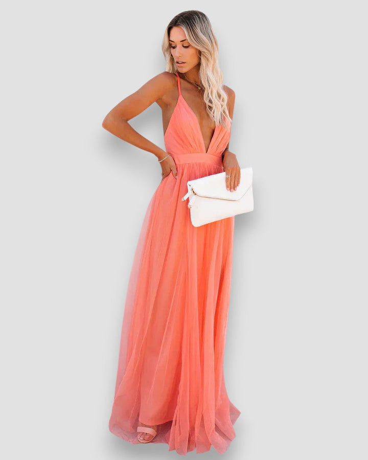 Model turned sideways, showcasing coral dress with V-neck and flowing skirt, holding white clutch, studio background.