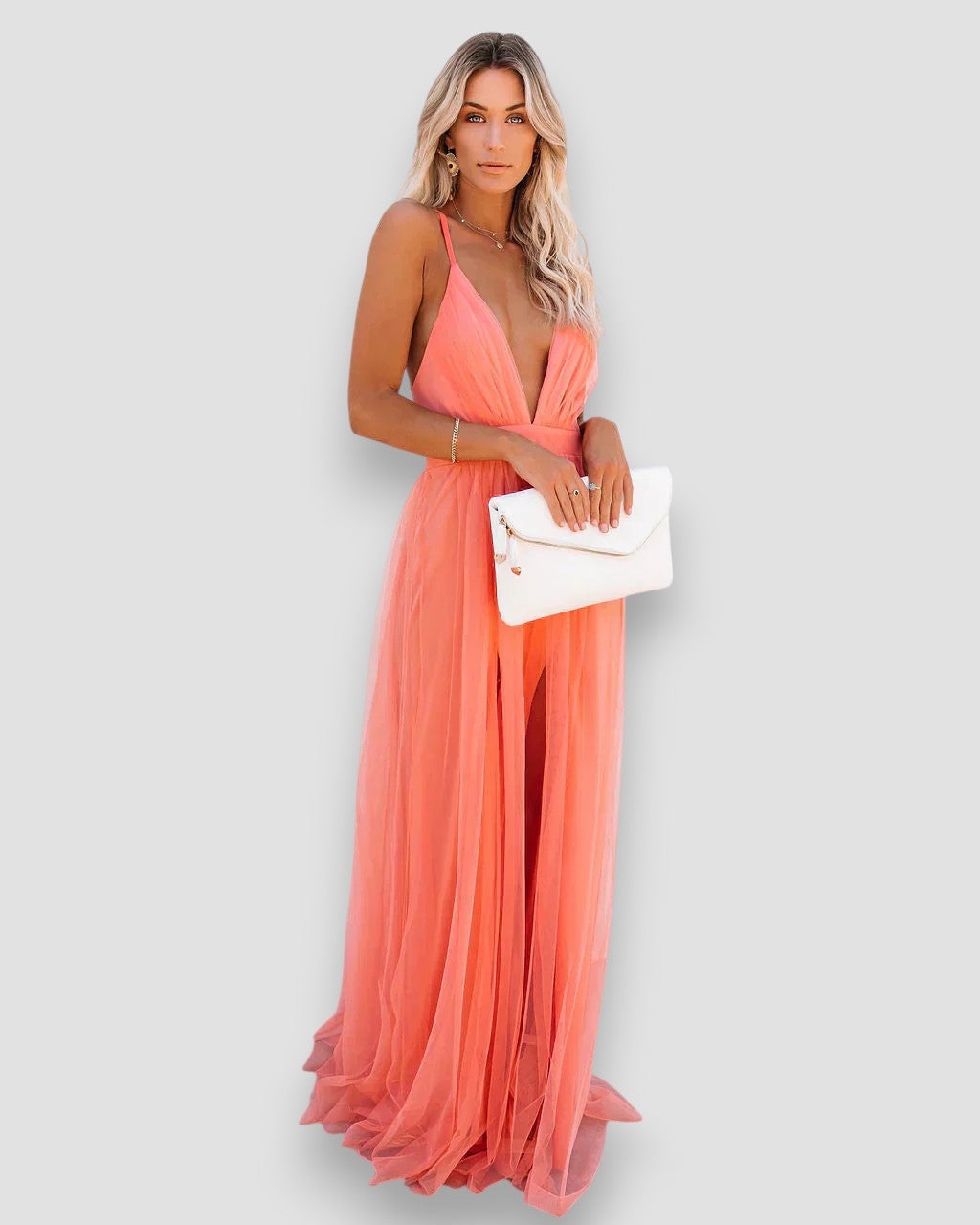 Model in a flowing coral dress with deep V-neck and spaghetti straps, holding a white clutch, studio background.