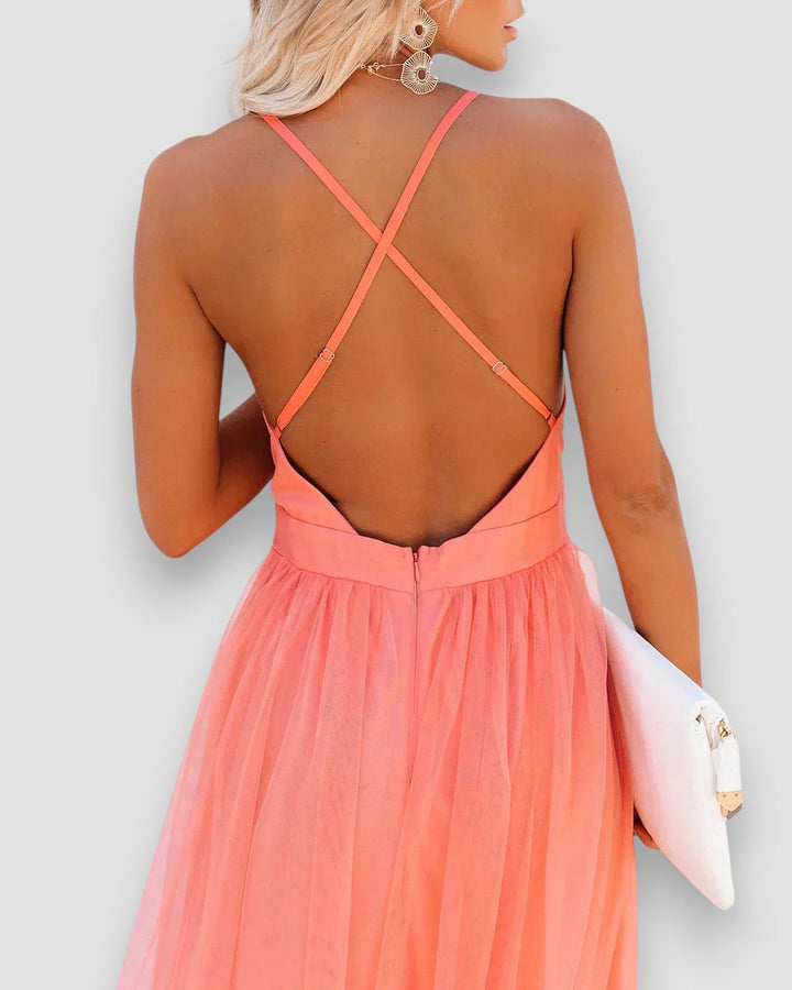 Back view of model in coral dress with crisscross straps, holding a white clutch, studio background.