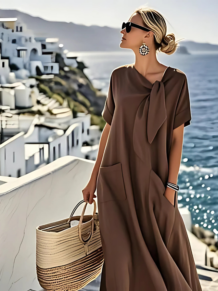 Elegant brown dress with pockets, worn by a woman with sunglasses and earrings, holding a woven tote. Coastal scenery in background.