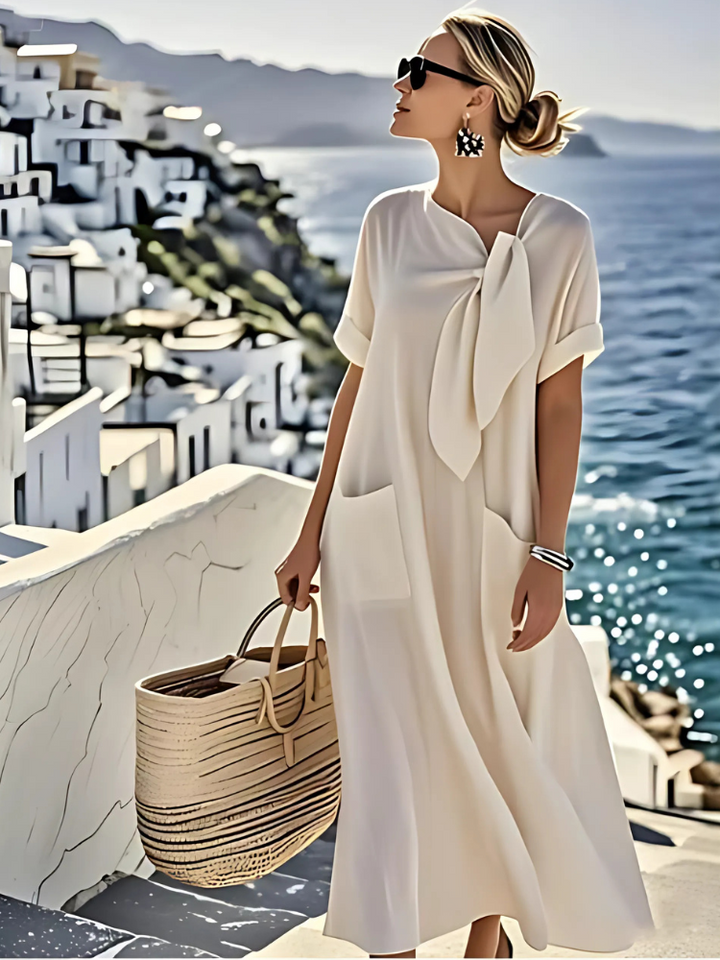 Elegant white dress with pockets, worn by a woman with sunglasses and earrings, holding a woven bag. Scenic coastal background.