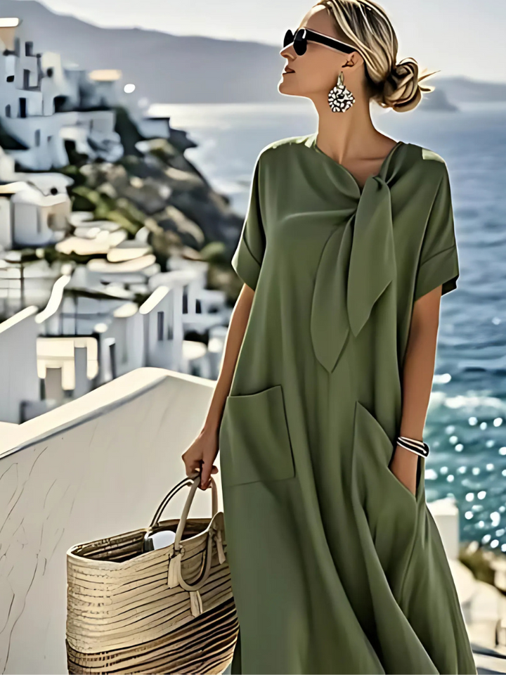 Elegant green dress with pockets, showcased by a woman wearing sunglasses and earrings, carrying a woven bag. Coastal view behind.