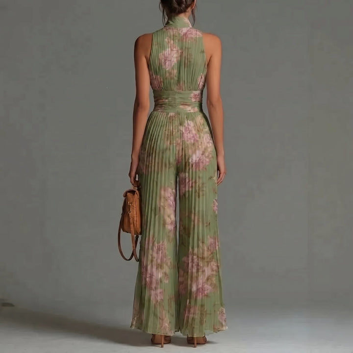 Back view of a model in green floral jumpsuit with pleated design, holding a brown bag, in a studio setting.