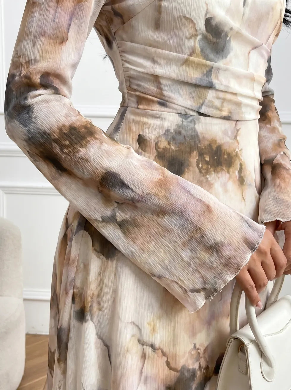 Close-up of a long-sleeve dress showing textured abstract watercolor pattern, hand holding a white handbag, indoor setting.