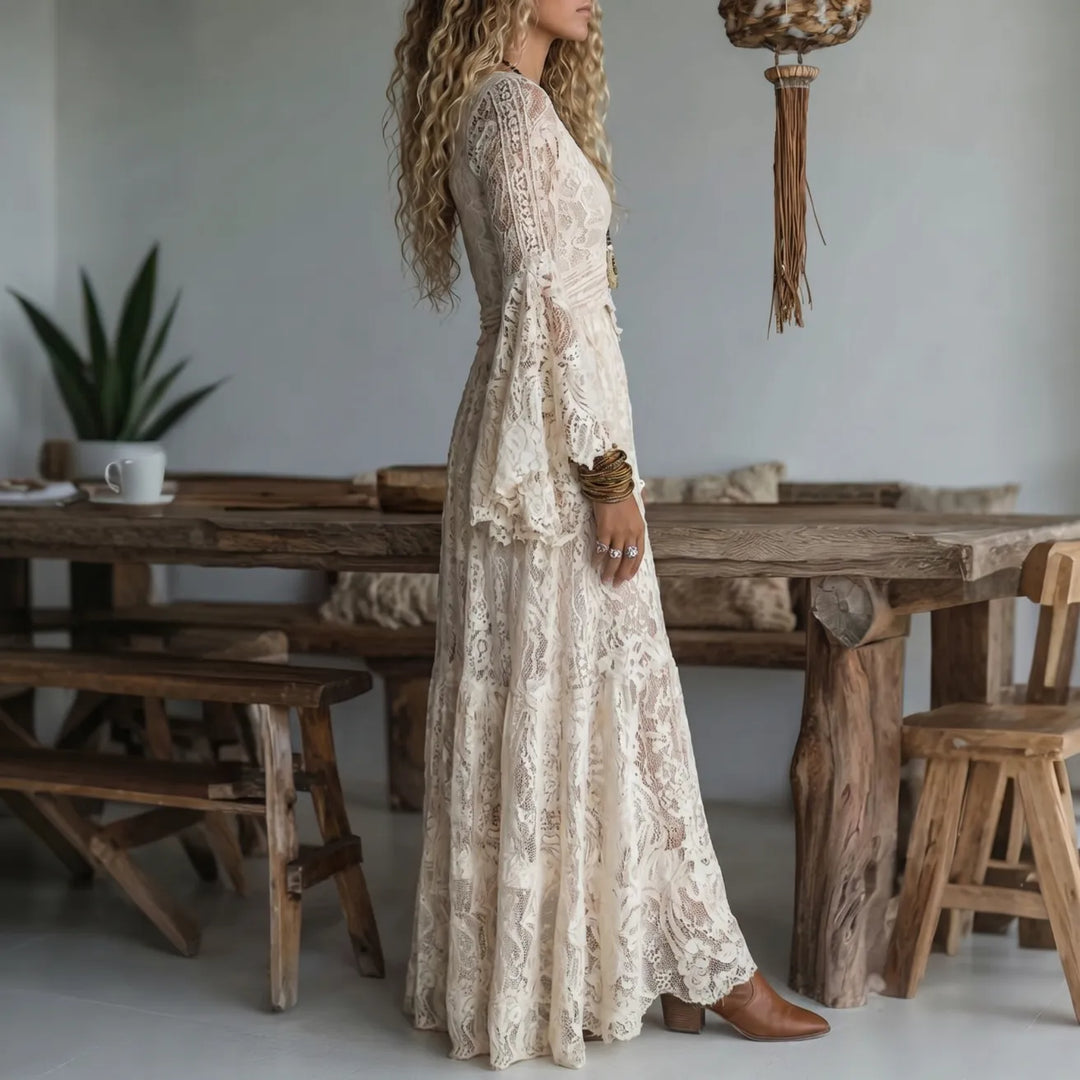 Side view of a woman in a cream lace maxi dress, in a rustic room with wooden dining set and earthy decor.