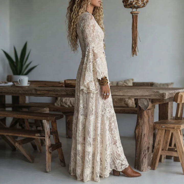 Side view of a woman in a cream lace maxi dress, in a rustic room with wooden dining set and earthy decor.
