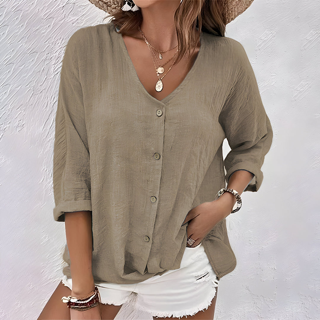 Woman in a beige relaxed button-up blouse with rolled sleeves, accessorized with jewelry, set against a textured white background.