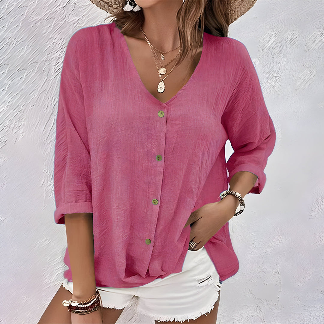 Model wearing a pink relaxed button-up blouse with rolled sleeves, paired with white shorts, against a textured white background.