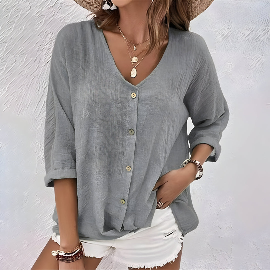 Woman wearing a gray relaxed button-up blouse with rolled sleeves, paired with white shorts, set against a textured white background.