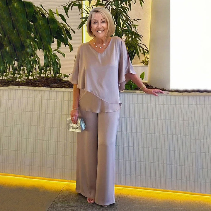 Woman in elegant beige two-piece set, in a modern indoor setting with plants and soft lighting.