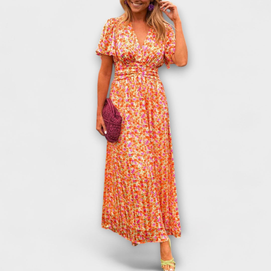 Woman wearing a colorful floral V-neck dress with short sleeves, holding a pink clutch, on a plain background.