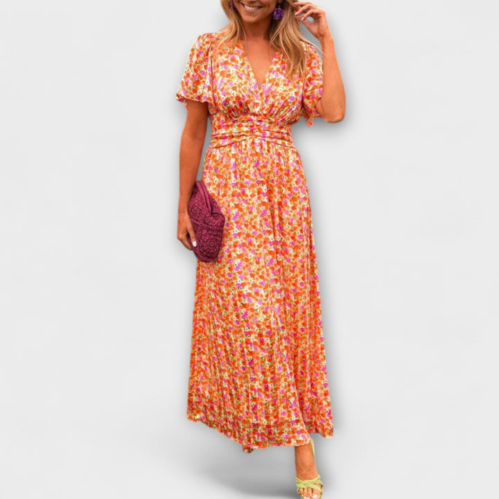 Woman wearing a colorful floral V-neck dress with short sleeves, holding a pink clutch, on a plain background.