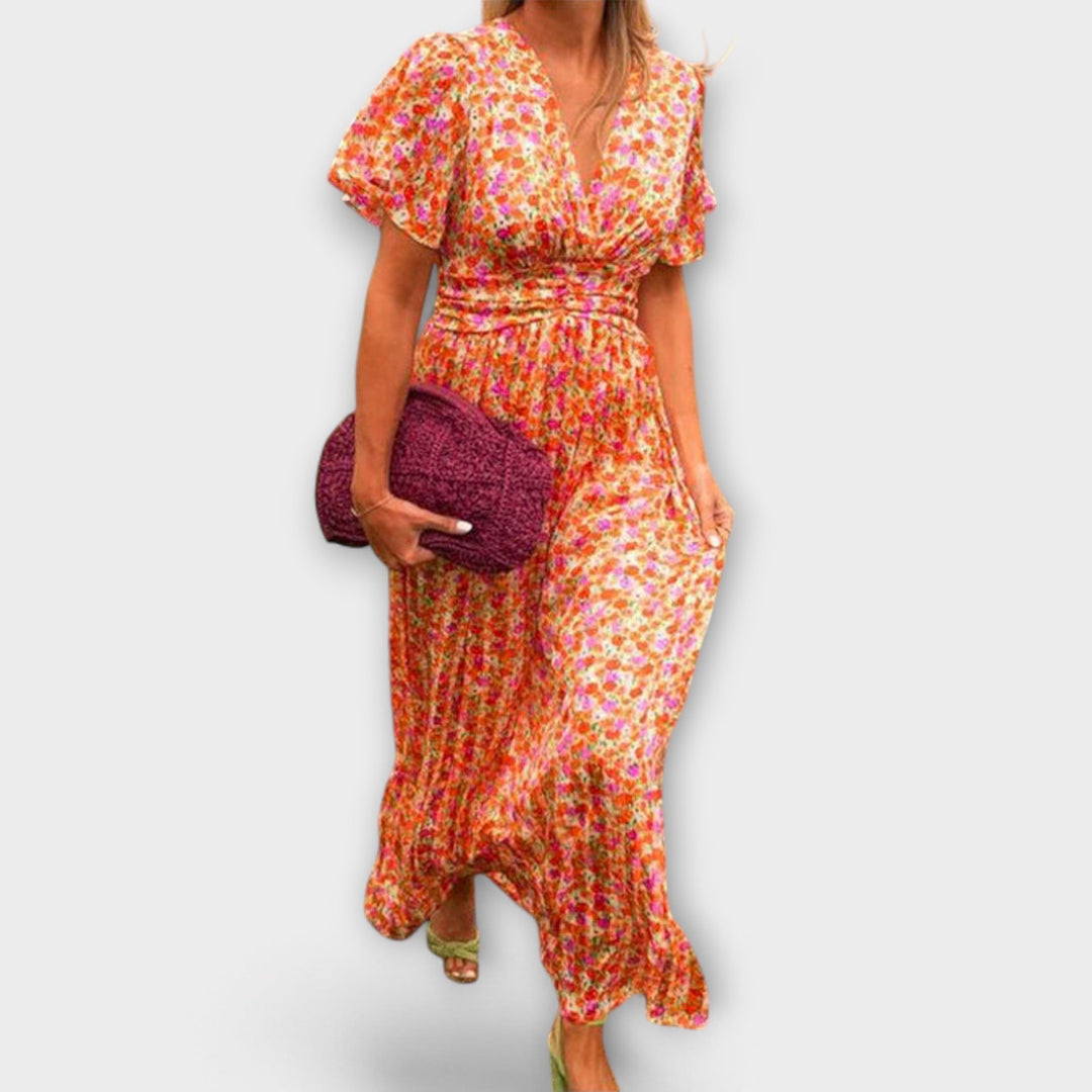 Woman walking in a floral V-neck dress with short sleeves, holding a pink clutch, on a plain backdrop.