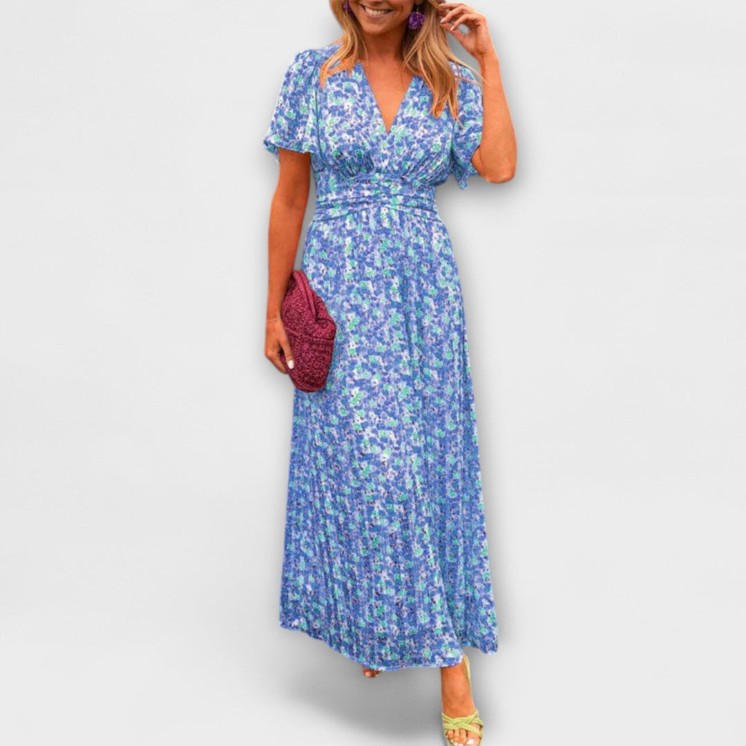 Model in a blue floral V-neck dress with short sleeves, holding a magenta clutch, on a plain backdrop.