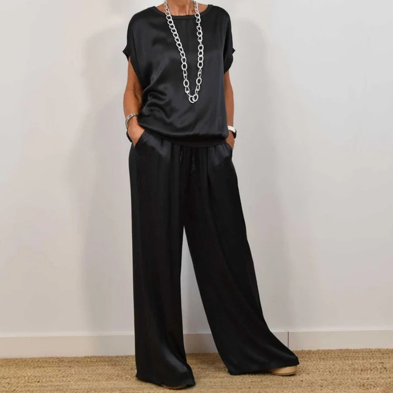 Sophisticated black two-piece set featuring a relaxed top and wide-leg pants, accessorized with a silver necklace, in a white studio.
