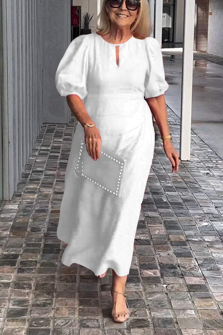Woman in white puff sleeve dress, holding clutch, walking on tiled path with modern gray wall backdrop.
