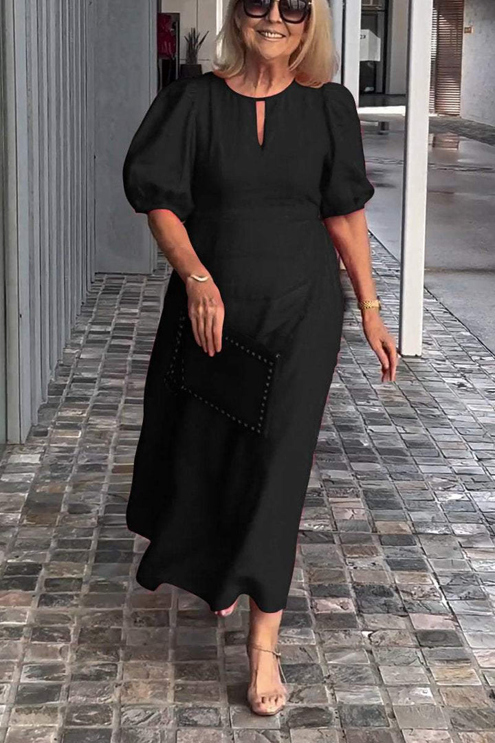 Woman in black puff sleeve dress, holding clutch, walking on tiled path with modern gray wall backdrop.