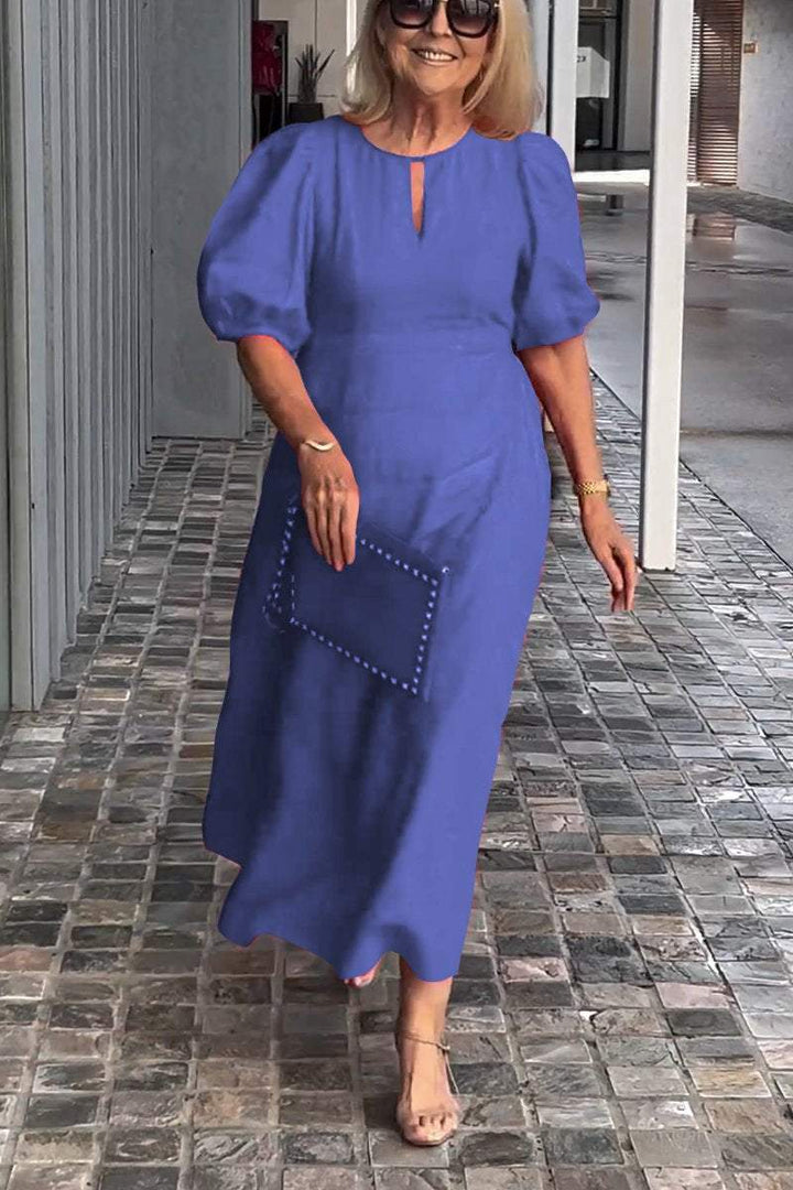 Woman in blue puff sleeve dress, holding clutch, walking on tiled path with modern gray wall backdrop.