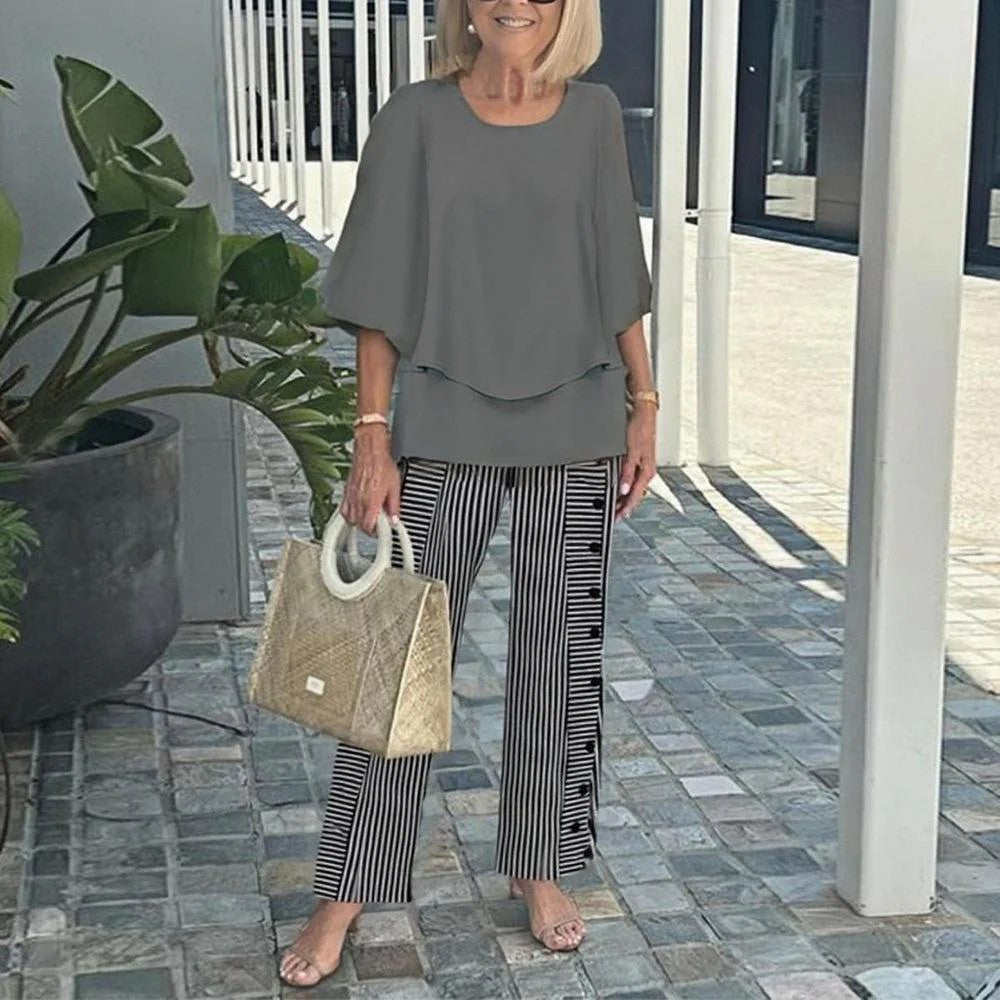 Woman wearing gray top and striped pants, holding a textured handbag, standing on a paved walkway with plants.