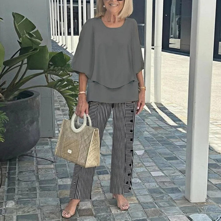 Woman wearing gray top and striped pants, holding a textured handbag, standing on a paved walkway with plants.