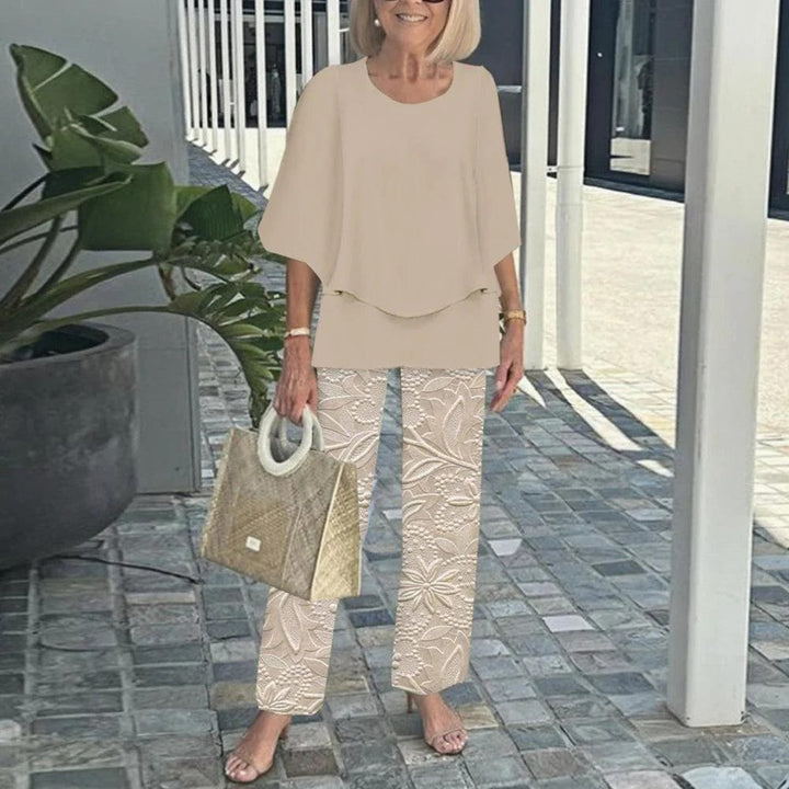 Woman in beige top and floral print pants, holding a textured handbag, on a paved walkway with plants.