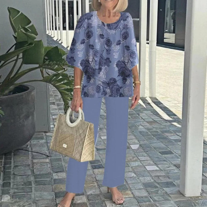 Woman in blue floral top with matching pants, holding a textured handbag, on a paved walkway with plants.