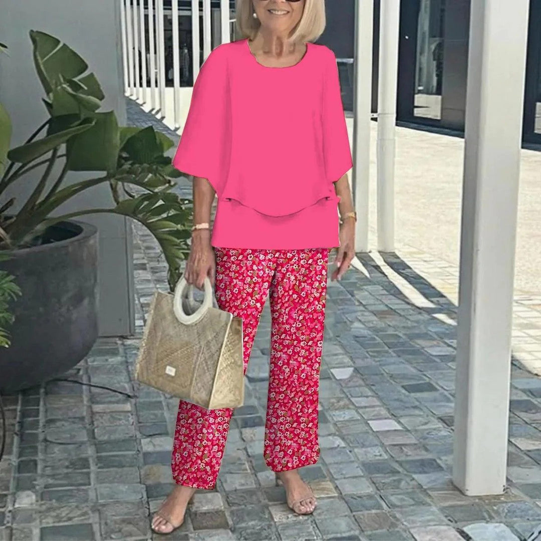 Woman in pink top with red floral pants, holding a textured handbag, on a paved walkway with plants.
