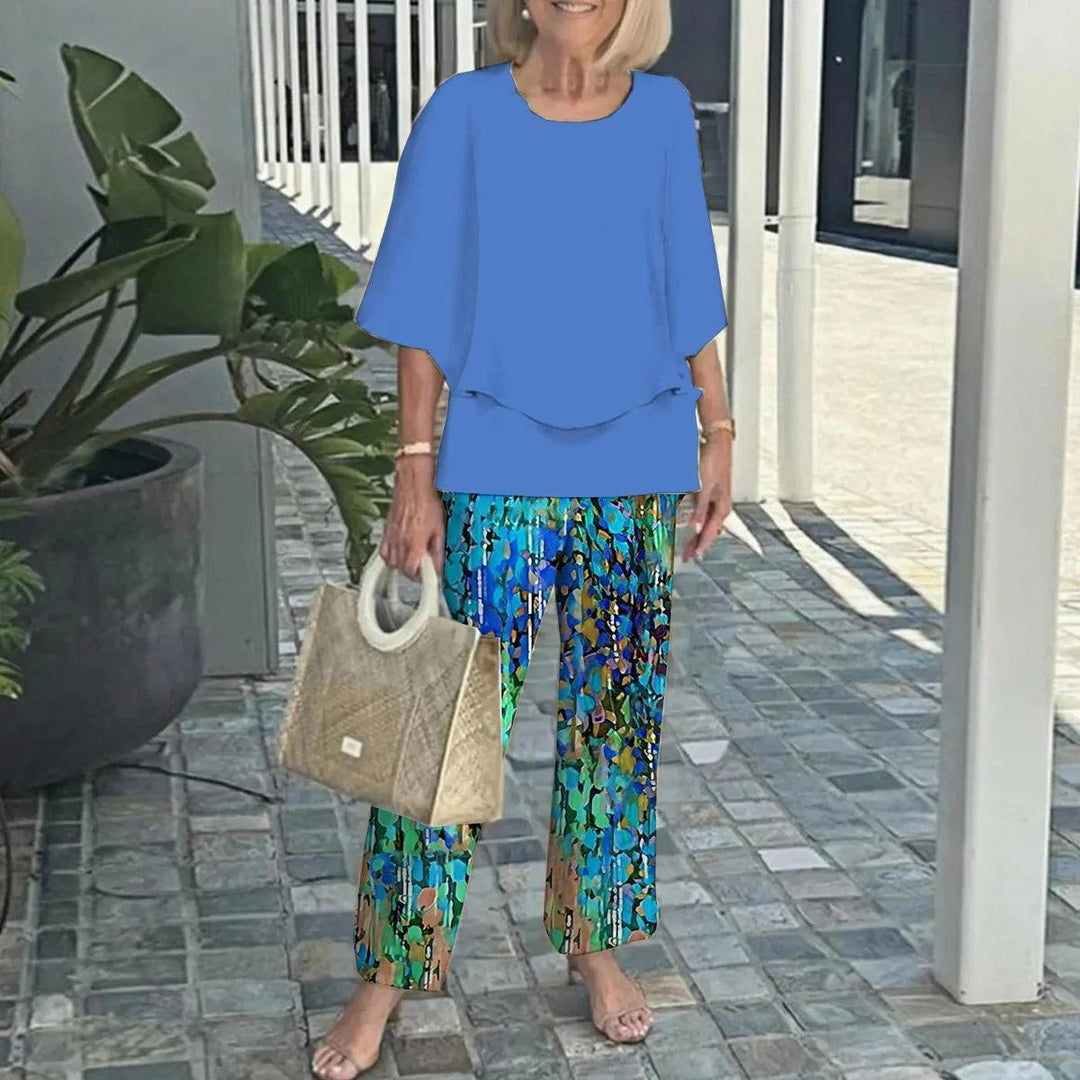 Woman in blue top and colorful abstract pants, holding a textured handbag, on a paved walkway with plants.