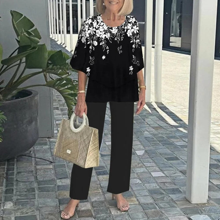 Woman in black floral top and matching pants, holding a textured handbag, on a paved walkway with plants.