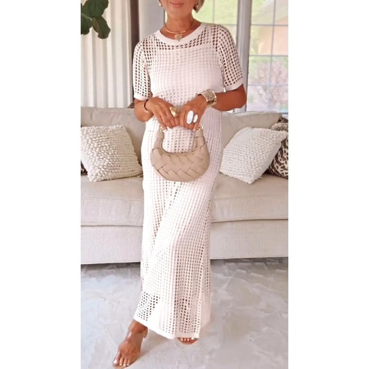 White cut-out maxi dress worn indoors, model holding a woven beige handbag, standing in front of a beige couch.