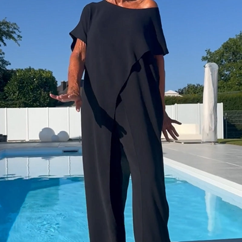 Elegant black off-shoulder two-piece set with draped detailing and loose pants, posed by a pool on a sunny day.