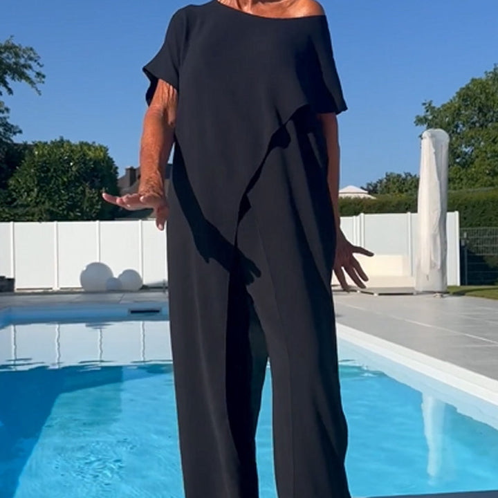 Elegant black off-shoulder two-piece set with draped detailing and loose pants, posed by a pool on a sunny day.
