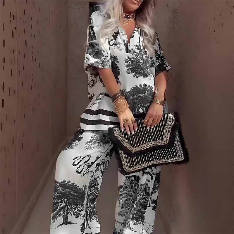 Person wearing a white and black tree-patterned V-neck top and pants, holding a fashionable clutch in a softly lit corridor.