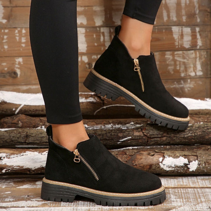 Side view of black ankle boots with zipper, worn with leggings against a wooden backdrop.