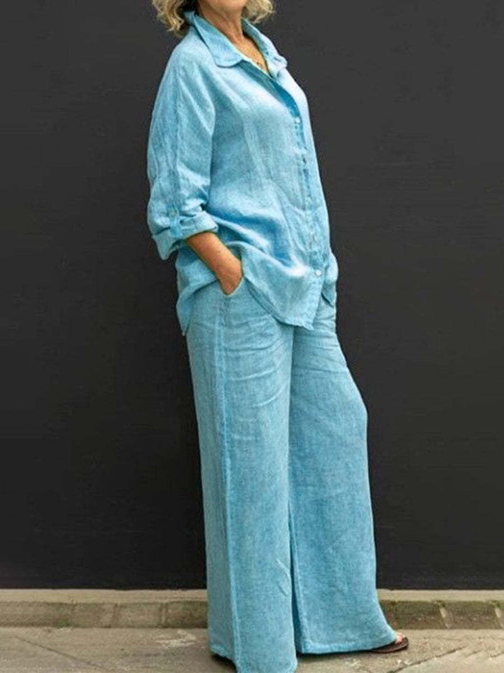 Blue effortless elegant set with long sleeves and wide-leg pants, worn by a woman against a black wall.