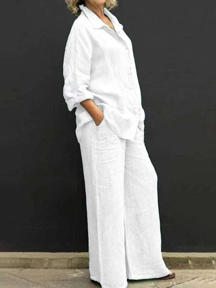 White effortless elegant set with long sleeves and wide-leg pants, worn by a woman against a black wall.