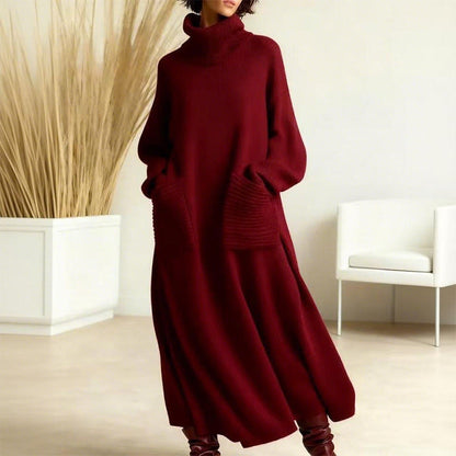 Elegant dark red sweater dress with a high neck and large pockets, modeled indoors with a minimalist beige interior and white chair.