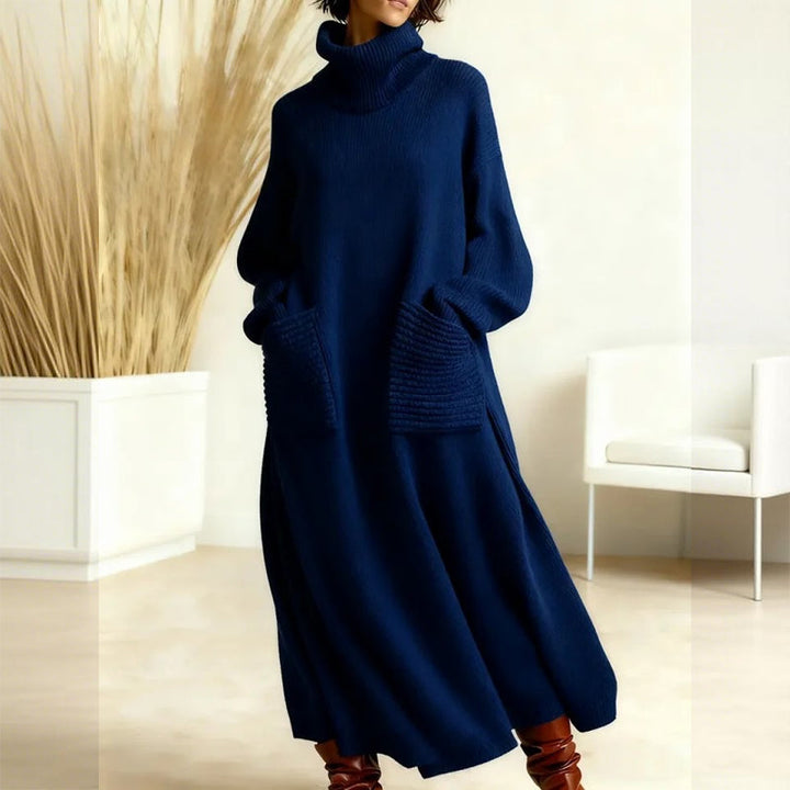 Elegant navy blue sweater dress with a high neck and large pockets, presented in a minimalist indoor setting with neutral decor and a white chair.