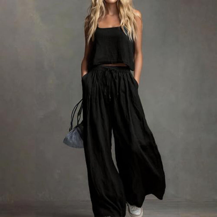 Woman wearing an elegant black set with a loose top and wide-leg pants, holding a small bag. Studio background.