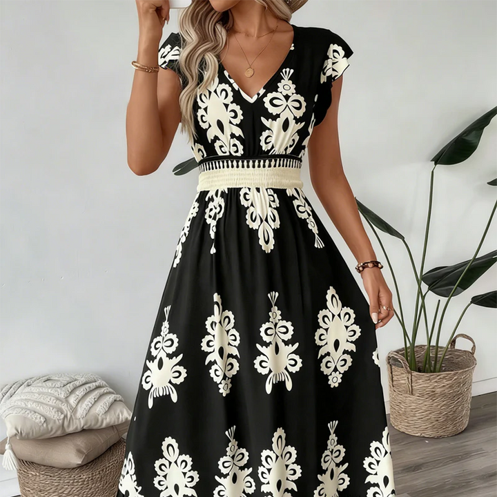 Black elegant long dress with white floral patterns and a smocked waist, worn indoors with plants and cushions.