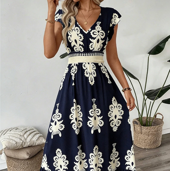 Navy blue elegant long dress with white floral patterns and a smocked waist, worn indoors with plants and cushions.