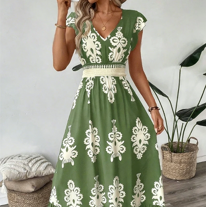 Green elegant long dress with white floral patterns and a smocked waist, worn indoors with plants and cushions.