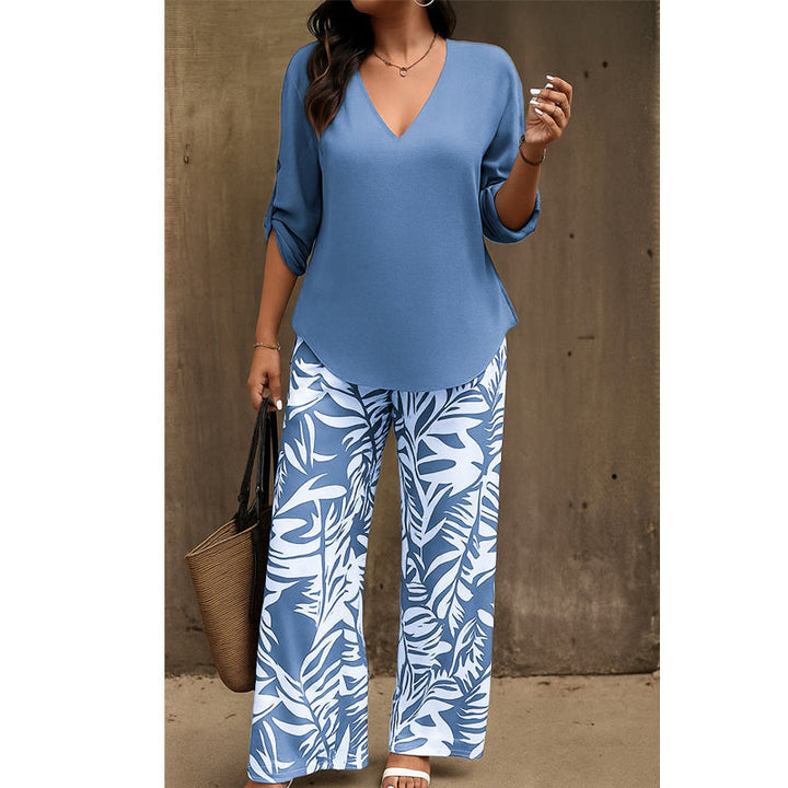 Woman in blue V-neck top and blue and white leaf-print pants, straw handbag in hand, standing near a textured wall.