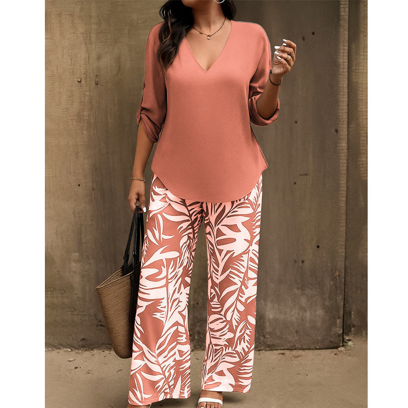 Woman in rust-colored V-neck top and rust-and-white leaf-print pants with a straw handbag against a rustic wall.